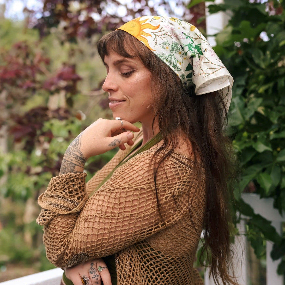 A woman wearing a brown crochet cardigan, olive green top, and a yellow and white patterned headscarf stands on a white porch with garden foliage in the background.