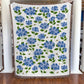 Floral-patterned blanket with blue flowers and green leaves on a wooden floor.