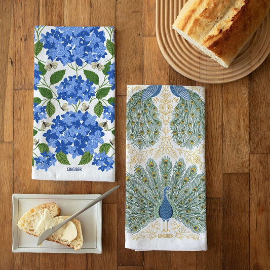 Two decorative tea towels with floral and peacock designs on a wooden surface with bread and butter.