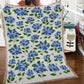 Floral-patterned throw blanket with blue flowers and green leaves on a wooden floor.