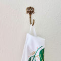 Decorative brass hook with a white towel featuring green leaf design on a light beige wall.