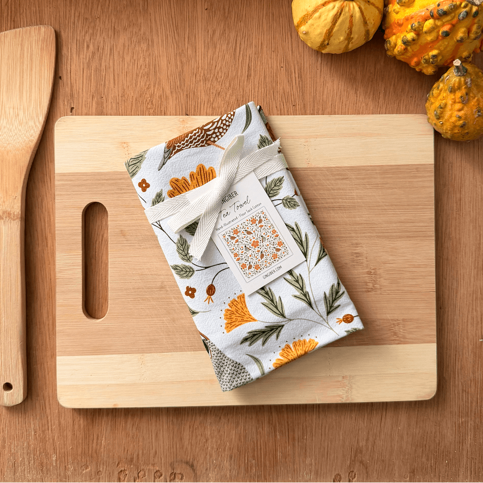 Floral-patterned towel on a wooden cutting board with pumpkins and a spatula.