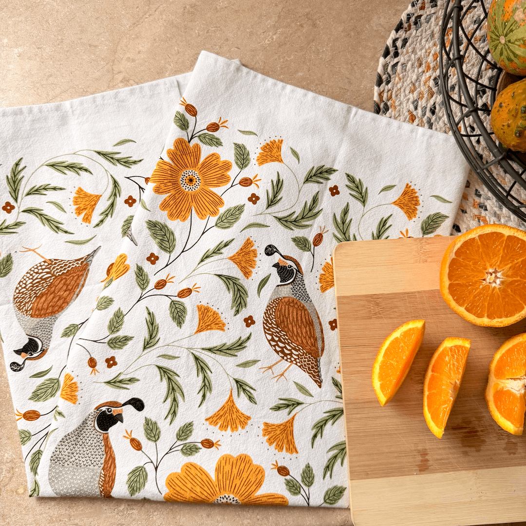 Decorative kitchen towel with floral and bird pattern on a wooden surface with oranges.