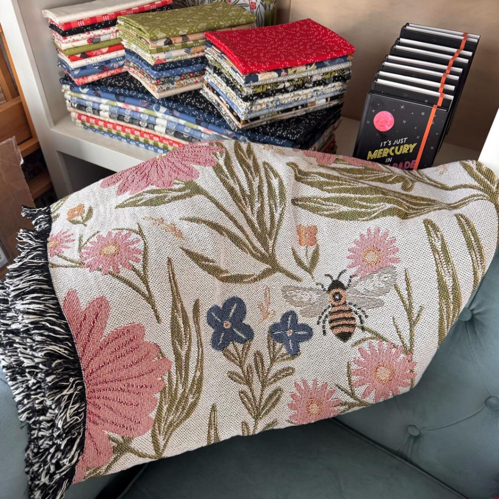 Floral and bee-patterned throw blanket draped over a couch with fabric stacks and books in the background.