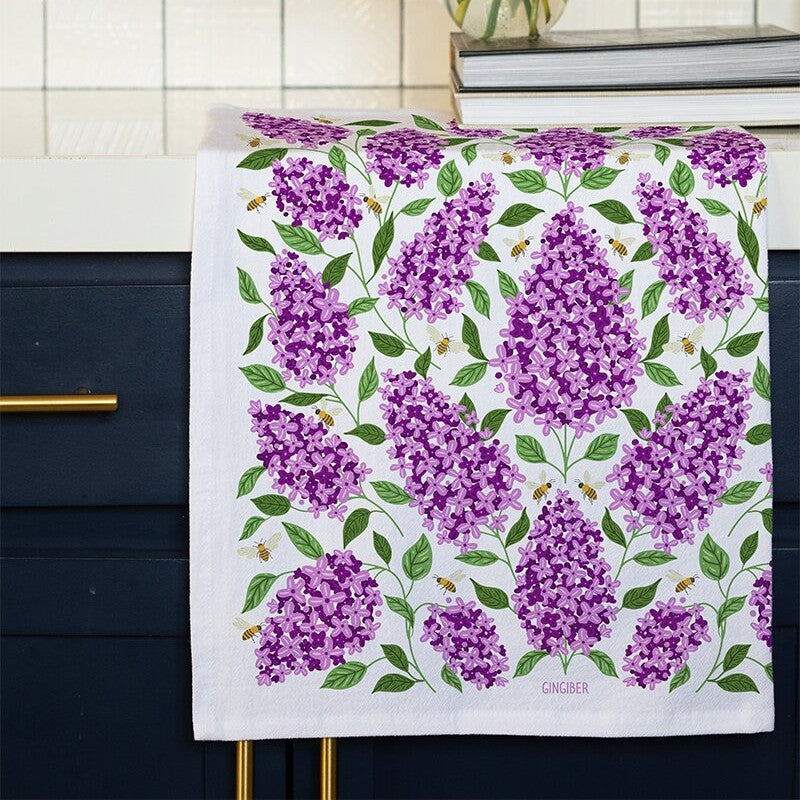 Tea towel with purple floral design hanging on a kitchen cabinet.