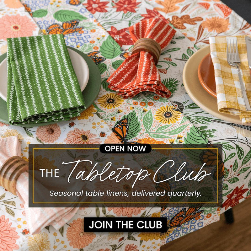 Colorful table setting with floral tablecloth, napkins, and cutlery, featuring The Tabletop Club advertisement.