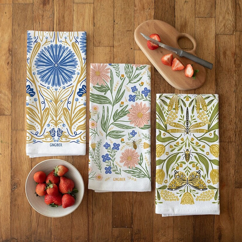 Three patterned towels on a wooden surface with strawberries and a cutting board.
