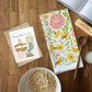 Floral towel with birds, card, bowl of oats, and wooden spoon on a wooden surface
