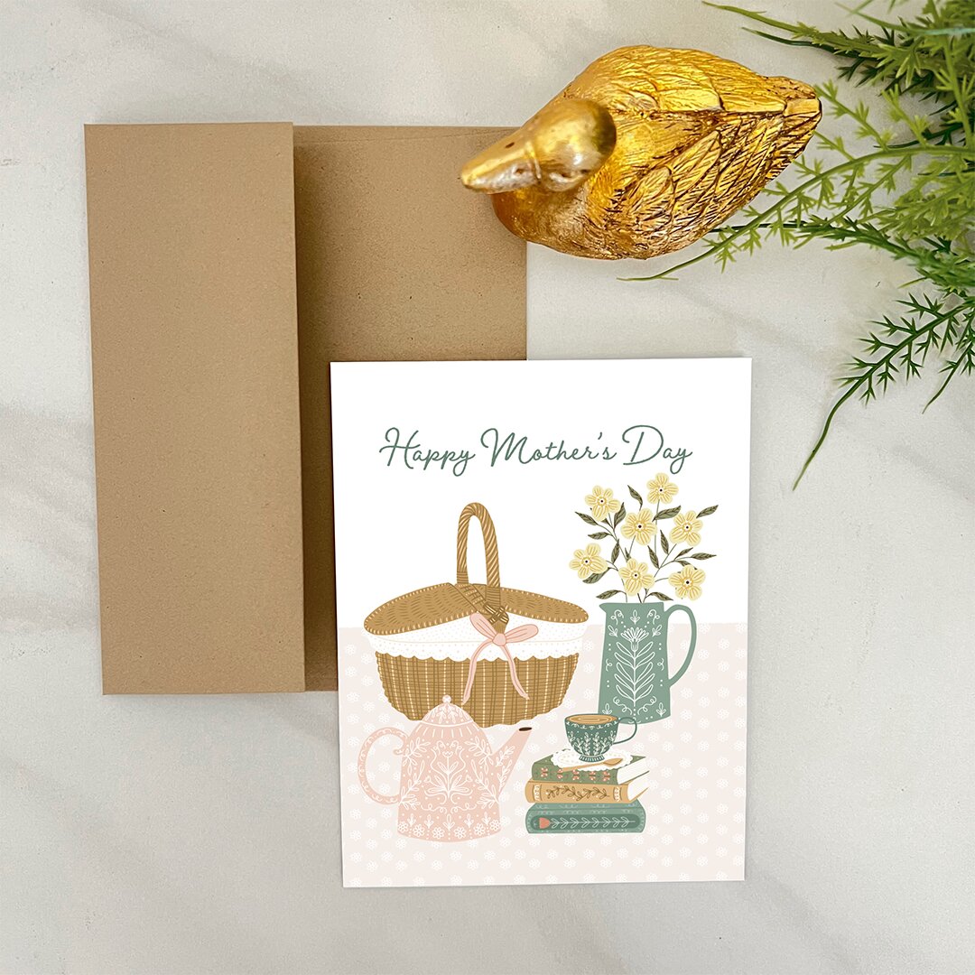Greeting card with 'Happy Mother's Day' text and illustrations of a teapot, basket, and flowers, accompanied by a gold duck and brown envelope on a light background.