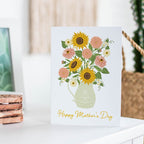 Greeting card with sunflowers and pink flowers in a pitcher on a white surface.