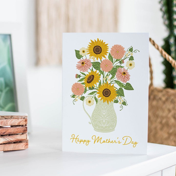 Greeting card with sunflowers and pink flowers in a pitcher on a white surface.