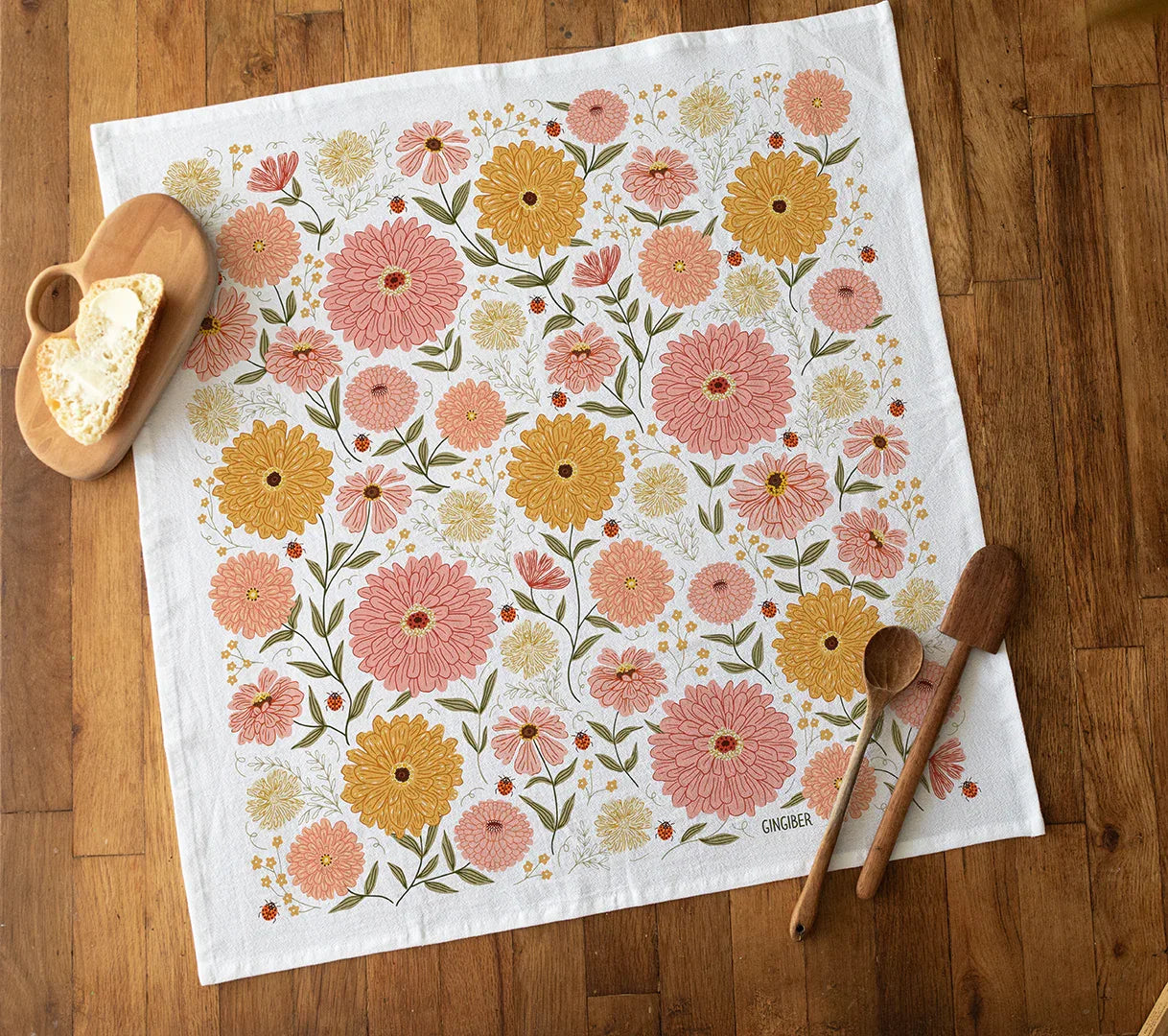Zinnia Apron and Tea Towel Bundle product image — features a floral tea towel design with pink and yellow zinnia patterns, kitchen setup with bread and utensils
